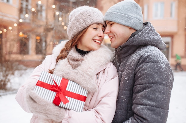 Feliz pareja amorosa afuera en el parque de invierno cubierto de nieve sosteniendo un regalo con cinta roja en las manos. Hombre y mujer alegres celebrando juntos el día de San Valentín. Cita en un día frío de invierno al aire libre.