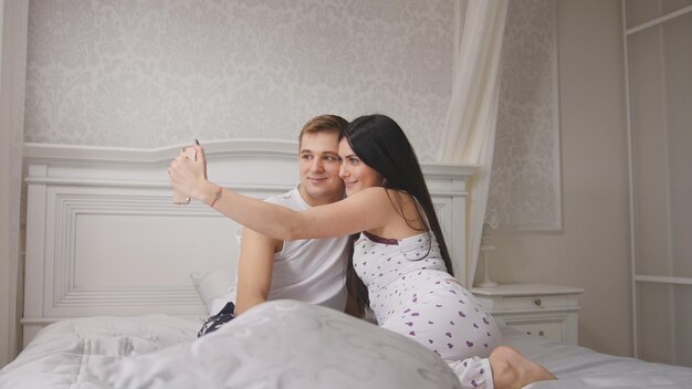 Feliz pareja amorosa alegre haciendo selfie en la cama joven atractivo chico y chica sentada en pijama