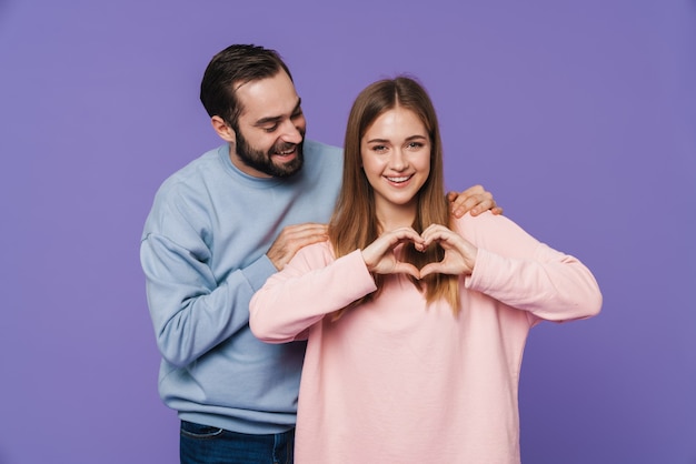 Feliz pareja amorosa hace gesto de amor de corazón aislado sobre pared púrpura.