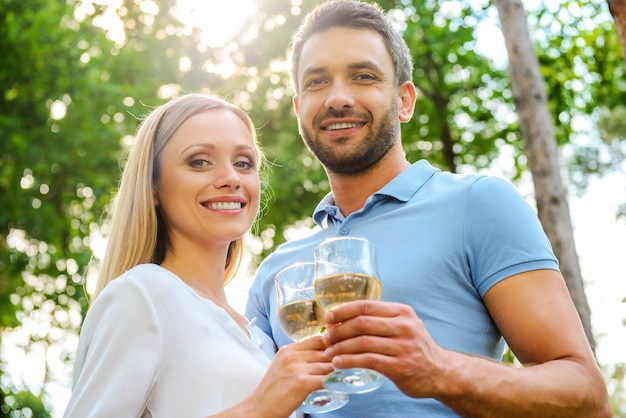 Feliz pareja amorosa. Vista de ángulo bajo de la feliz pareja amorosa joven sosteniendo vasos con vino blanco y sonriendo mientras está de pie cerca uno del otro al aire libre