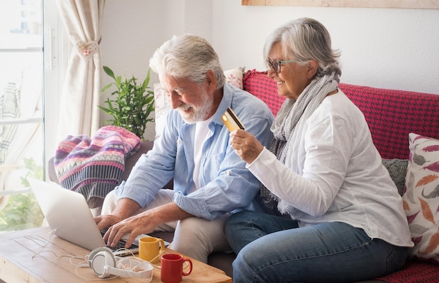 Feliz pareja de ancianos de los 70 sentada en un sofá con una computadora portátil, usa internet y tarjeta de crédito para comprar en línea