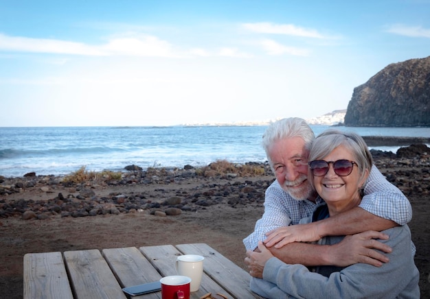 Una feliz pareja de ancianos abrazándose en la playa Sentado en una mesa de madera con dos tazas de café Dos personas reales