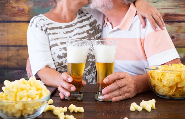 Feliz pareja de ancianos se besan mientras se sientan en una mesa de madera brindando con dos vasos de cerveza y papas fritas
