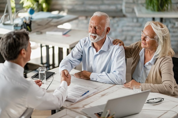 Feliz pareja de ancianos dándose la mano con un agente de seguros después de un acuerdo exitoso sobre una reunión en la oficina