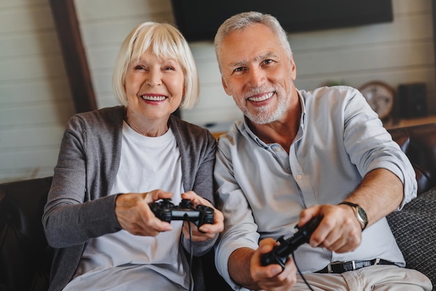 Feliz pareja de ancianos sentados juntos en un sofá sosteniendo controladores mientras juegan videojuegos en casa