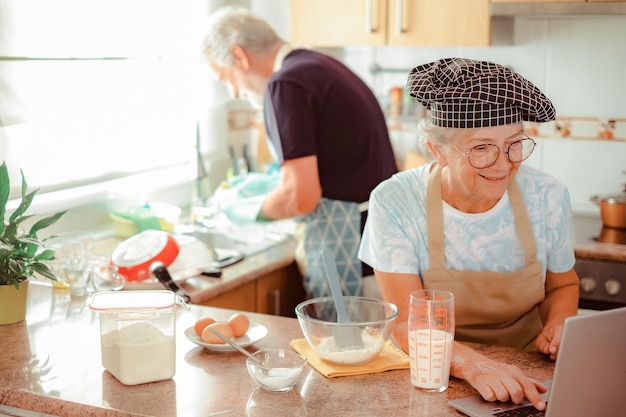 Feliz pareja de ancianos trabajando juntos en la cocina de la casa esposo lavando platos esposa preparando un pastel casero mirando páginas web por computadora portátil para la receta correcta