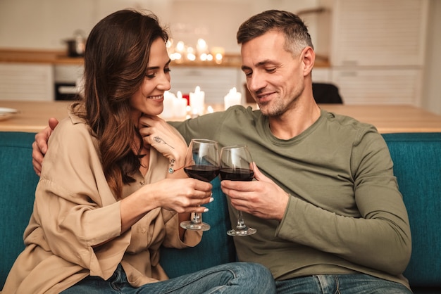 Feliz pareja apasionada bebiendo vino tinto de vasos mientras tiene una cena romántica a la luz de las velas en casa