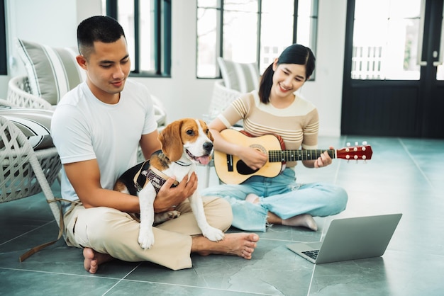 Feliz pareja asiática de adultos jóvenes trabajando en línea en casa y jugando con un perro beagle mientras usa una computadora portátil en la sala de estar