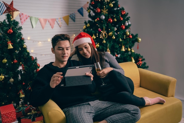 Feliz pareja asiática brindando champán y viendo películas juntos en una tableta sentados en el sofá con un árbol de Navidad y luces en el fondo Novio y novia en la noche de Nochebuena