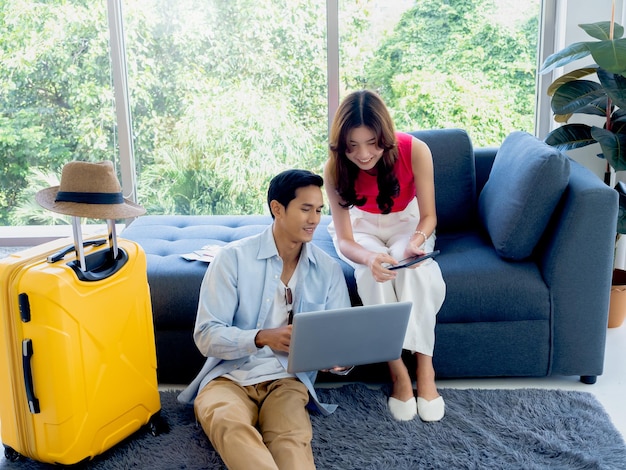 Feliz pareja asiática hombre sentado en el piso y mujer joven en el sofá mirando y usando computadora portátil y tableta juntos para reservar vuelos e información de viaje en la sala de estar viajes vacaciones de verano