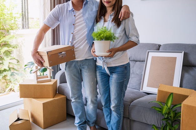 Feliz pareja asiática joven en sala de estar en casa nueva con pila de cajas de cartón en día de mudanza