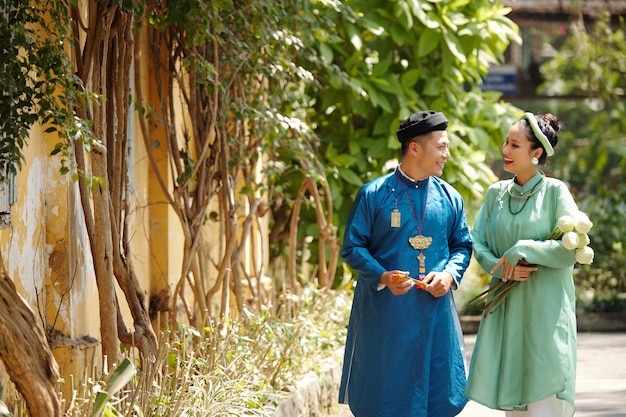 Feliz pareja asiática joven en vestidos tradicionales y piezas para la cabeza riendo y hablando al caminar al aire libre en un día de verano