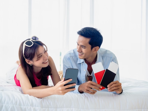 Feliz pareja asiática con reserva de vuelo para un viaje de verano al extranjero Un hombre y una mujer jóvenes con un teléfono inteligente casual y dos pasaportes y boletos mientras están acostados en la cama juntos se miran el uno al otro