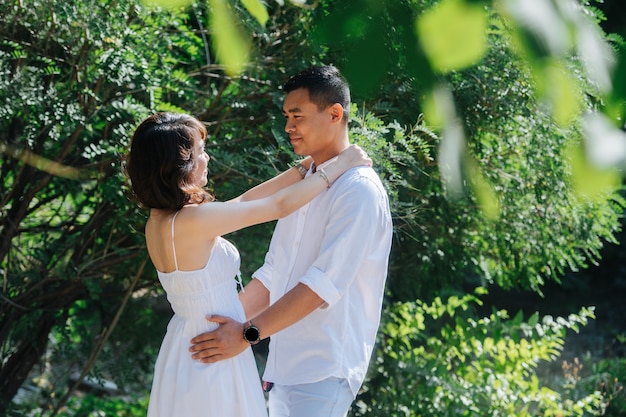 Feliz pareja asiática vestida de blanco de pie en un arbusto en un parque, lejos de los ojos de los demás. Mirándose, abrazándose.