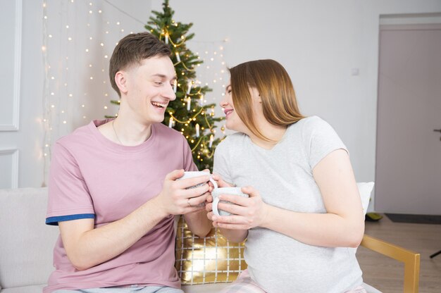 Feliz pareja bebiendo té o café en casa por la noche sobre las luces del árbol de Navidad en el fondo. Concepto de vacaciones, ocio y personas.