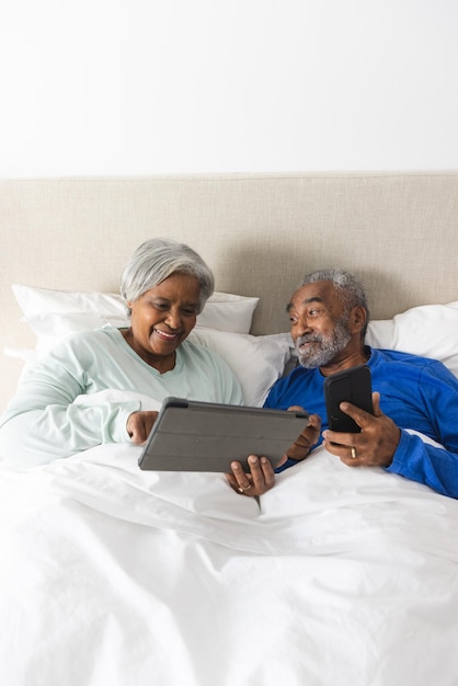 Una feliz pareja biracial de ancianos acostada en la cama y usando tableta y teléfono inteligente en casa