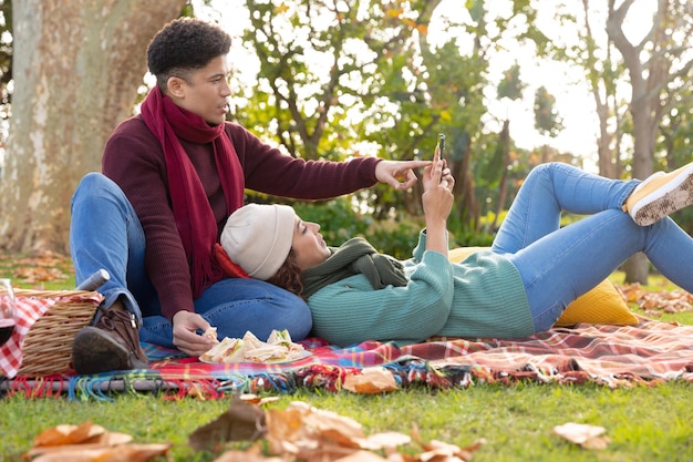 Feliz pareja birracial haciendo un picnic en una alfombra y usando un teléfono inteligente en el jardín de otoño, sonriendo. Concepto de inclusión, vida doméstica, tiempo libre, romance y convivencia.