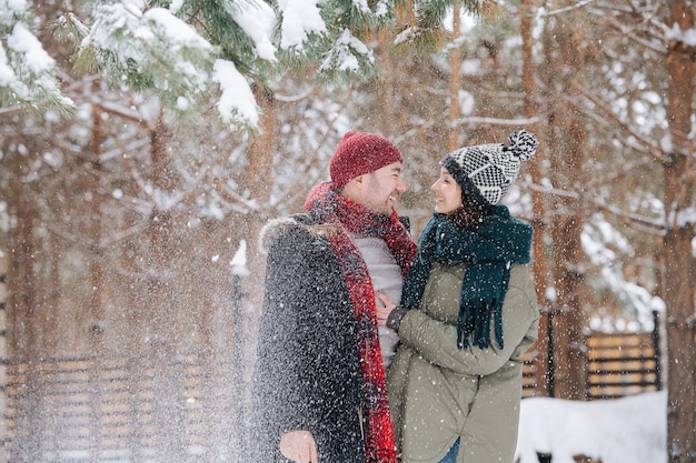 Feliz pareja caminando cerca uno del otro entre coníferas nevadas en el parque. Ambos con sombreros, chaquetas y bufandas. Disparo a través de partículas de nieve que caen de las ramas.