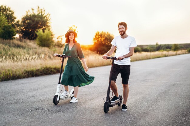 Feliz pareja caminando en scooters. Una mujer joven con vestido y sombrero en puesta de sol