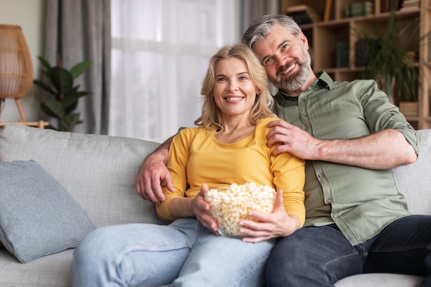 Feliz pareja casada de mediana edad viendo televisión y comiendo palomitas de maíz en casa