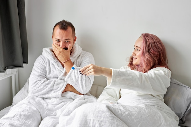 Feliz pareja caucásica emocionada con prueba de embarazo positiva en la cama por la mañana, reacción feliz, el hombre está sorprendido