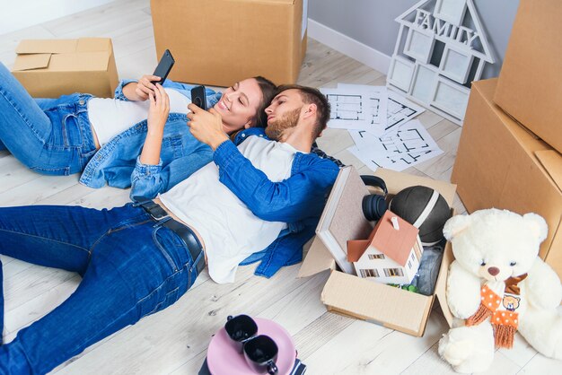 Feliz pareja caucásica joven acostado en el piso de madera y mensajes en teléfonos durante tener descanso mientras se muda a nueva casa.