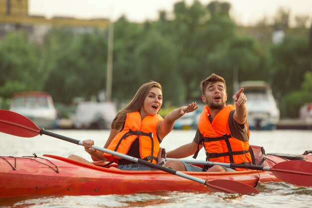 Feliz pareja caucásica joven kayak en el río con puesta de sol en los fondos. Divirtiéndose en la actividad de ocio. Modelo masculino y femenino feliz riendo en el kayak. Deporte, concepto de relaciones. Vistoso.