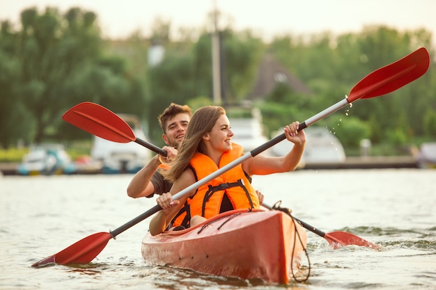 Feliz pareja caucásica joven kayak en el río con puesta de sol en los fondos. Divirtiéndose en la actividad de ocio. Modelo masculino y femenino feliz riendo en el kayak. Deporte, concepto de relaciones. Vistoso.