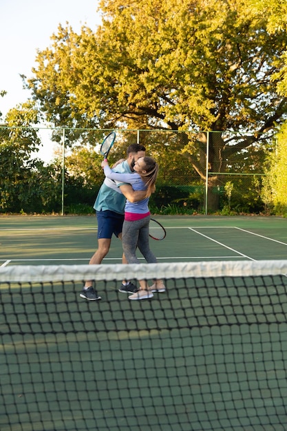 Feliz pareja caucásica jugando al tenis abrazándose en la cancha de tenis al aire libre