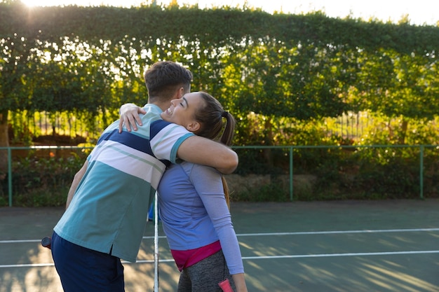 Feliz pareja caucásica jugando al tenis abrazándose a través de la red en la cancha de tenis al aire libre