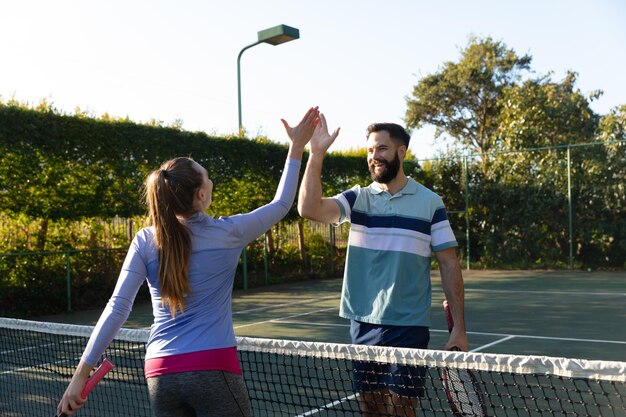 Feliz pareja caucásica jugando tenis estrechando la mano sobre la red en la cancha de tenis al aire libre