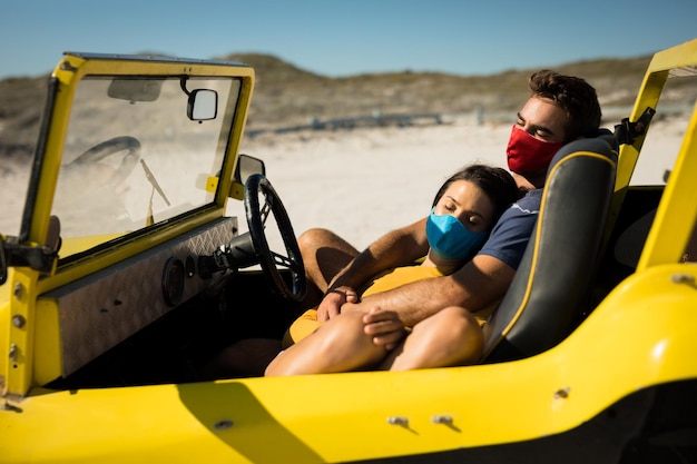Feliz pareja caucásica con máscaras faciales sentadas en un buggy de playa relajándose. parada en la playa en un viaje de vacaciones de verano durante la pandemia de coronavirus covid 19.