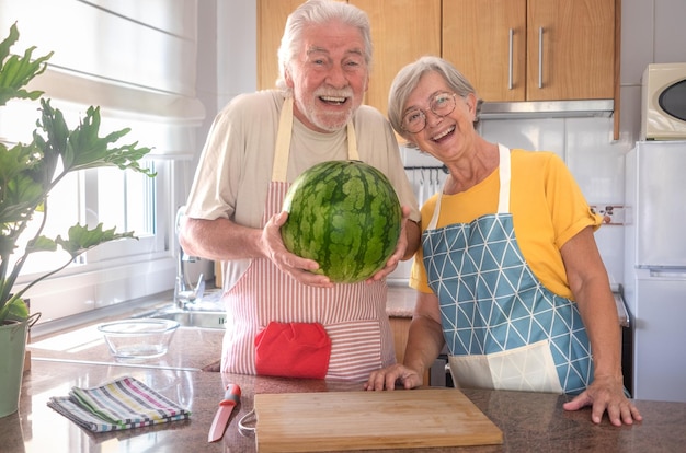 Feliz pareja caucásica senior en la cocina de casa sosteniendo una sandía grande y pesada mirando a la cámara sonriendo hidratación dieta de frescura y concepto de estilo de vida de alimentación saludable