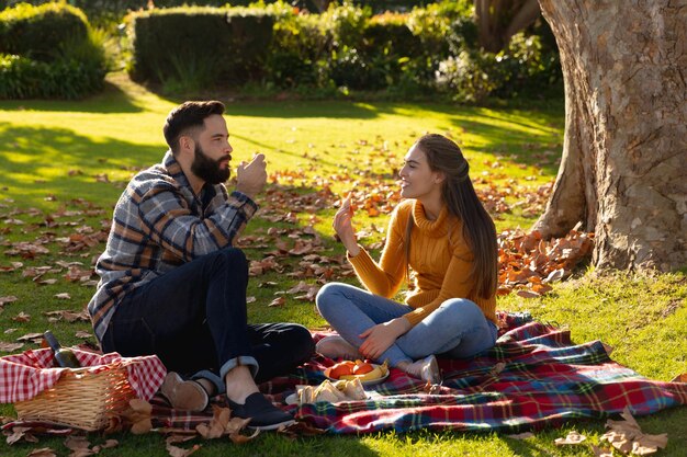 Feliz pareja caucásica sentada en una alfombra haciendo un picnic y hablando en el soleado jardín de otoño. Concepto de vida doméstica, tiempo libre, romance y convivencia.