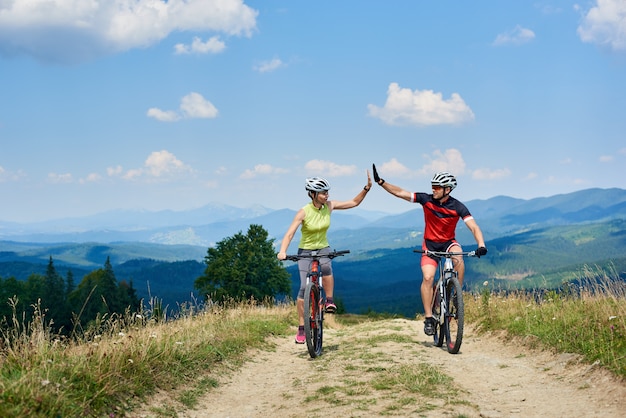 Feliz pareja ciclistas andar en bicicleta a campo traviesa en la carretera de montaña en un día soleado de verano en los Cárpatos. Hombre y mujer activos que se dan unos cinco