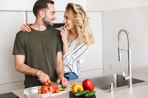 Feliz pareja cocinando juntos mientras está de pie en la cocina, cortando verduras a bordo, abrazando