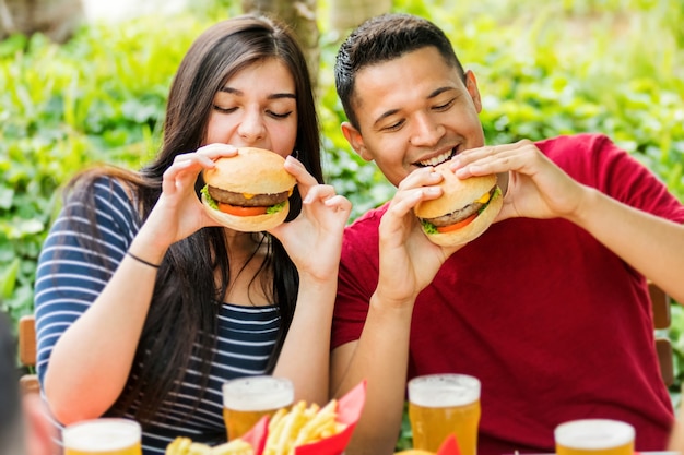 Foto feliz pareja comiendo hamburguesas y bebiendo cervezas heladas en un restaurante al aire libre en una vista de cerca de ellos sonriendo con anticipación mientras muerden la comida