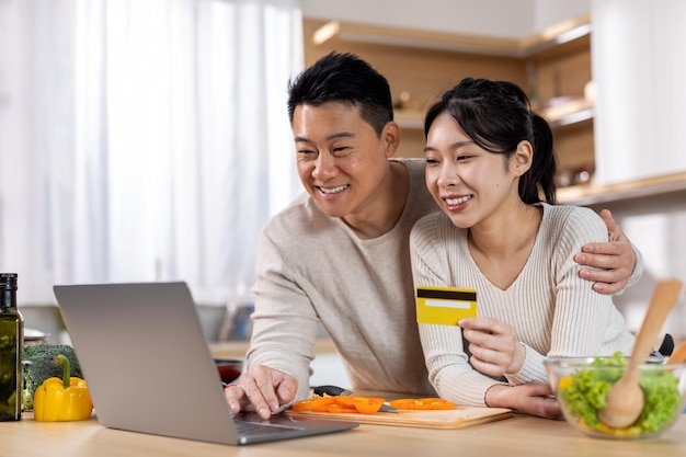 Feliz pareja coreana haciendo un pedido en línea usando una computadora portátil en la cocina
