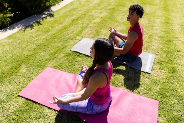 Feliz pareja diversa en forma practicando meditación de yoga sentada en un jardín soleado. Verano, estilo de vida saludable, relajación y vacaciones.