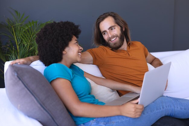 Feliz pareja diversa sentada en un sofá abrazándose y usando una computadora portátil. permanecer en casa aislado durante el cierre de cuarentena.