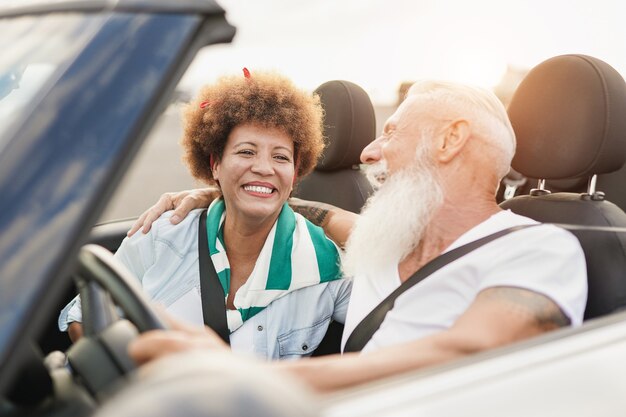 Feliz pareja divirtiéndose dentro de un coche convertible - Personas mayores multirraciales en un viaje por carretera con coche descapotable