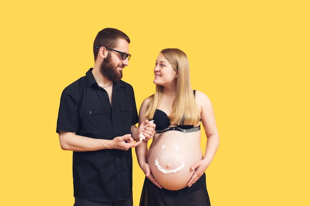 Feliz pareja embarazada con emoji en el vientre sobre fondo amarillo