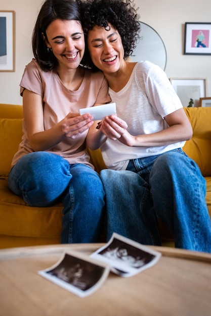 Feliz pareja embarazada lesbiana multirracial mirando los primeros ultrasonidos de bebé en casa Imagen vertical