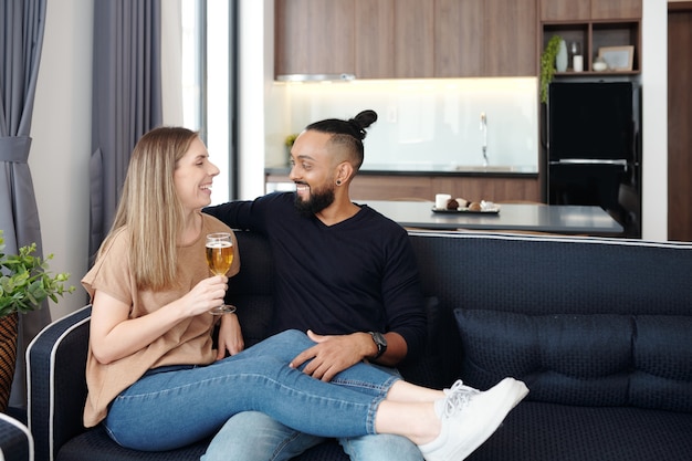 Feliz pareja de enamorados descansando en el sofá en casa, bebiendo vino, bromeando y riendo