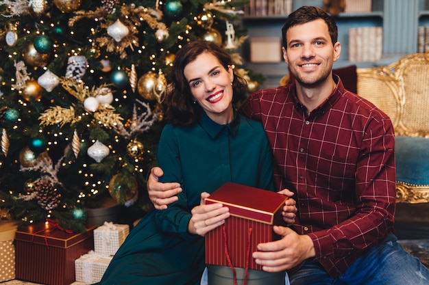 Feliz pareja encantadora en el amor, abrazarse, sentarse cerca del árbol de Año Nuevo decorado, mantener presente