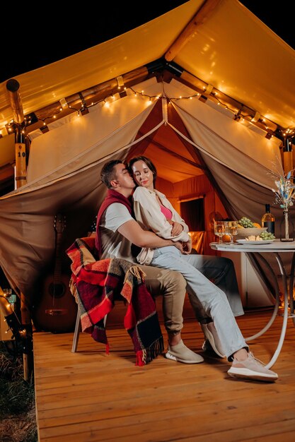 Feliz pareja encantadora relajándose en glamping en las noches de verano y bebiendo vino cerca de una acogedora hoguera Tienda de campaña de lujo para recreación al aire libre y recreación Concepto de estilo de vida