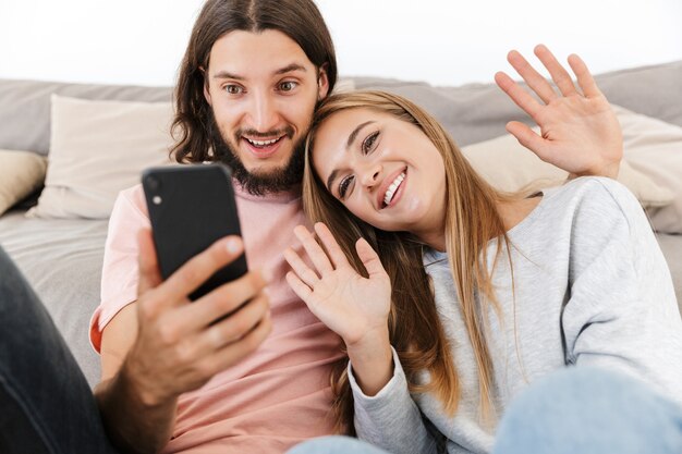 Feliz pareja encantadora relajándose en un sofá en casa, teniendo una videollamada mientras sostiene el teléfono móvil, agitando las manos