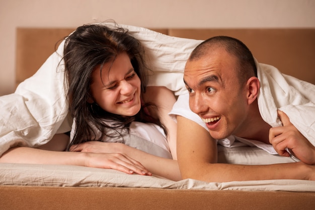 Feliz pareja se encuentran en una cama cubierta con una manta sobre su cabeza.