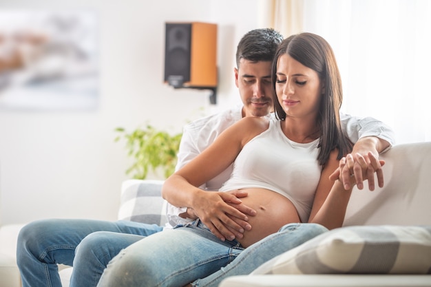 Feliz pareja esperando un bebé se sienta en un sofá, con ambas manos en el vientre.