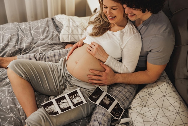 Feliz pareja esperando mirando el examen médico de su bebé mientras se relaja en una cama en el dormitorio.